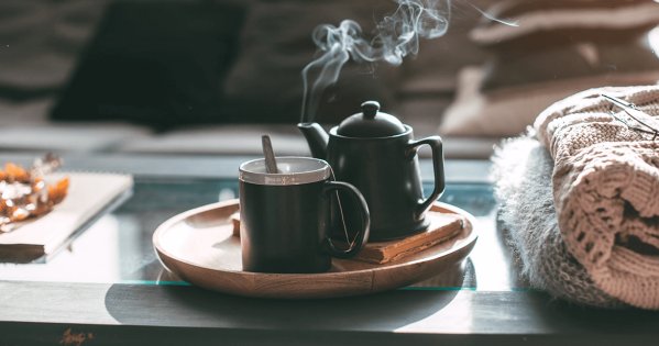 Heiße Tasse Kaffe mit einer Kanne auf einen Holzbrett daneben ein Buch und eine Wolldecke