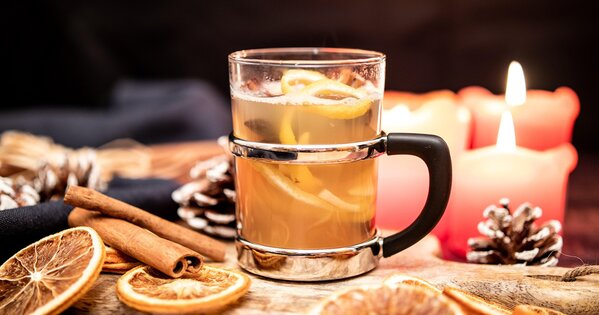 Ein weihnachtlich dekorierter Tisch, in der Mitte ein Becher mit Orangenscheiben und Punch. rechts rot-orangene Kerzen, davor ein Tannenzapfen. links Zimtstangen, Orangenscheiben und eine Serviertte in blau.